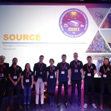 Gruppenbild des KSat Teams beim Fly Your Satellite Auswahlworkshop vor einer großen Projektorleinwand, auf der eine SOURCE Powerpointfolie abgebildet ist